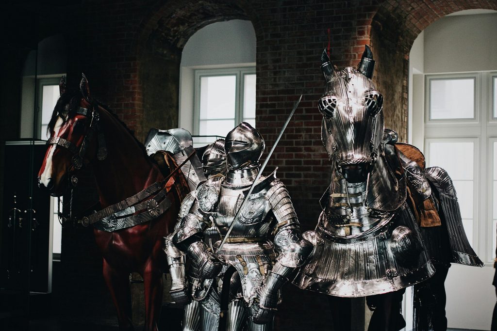 Medieval knight in full armor with a swordsman and armored horse indoors.
